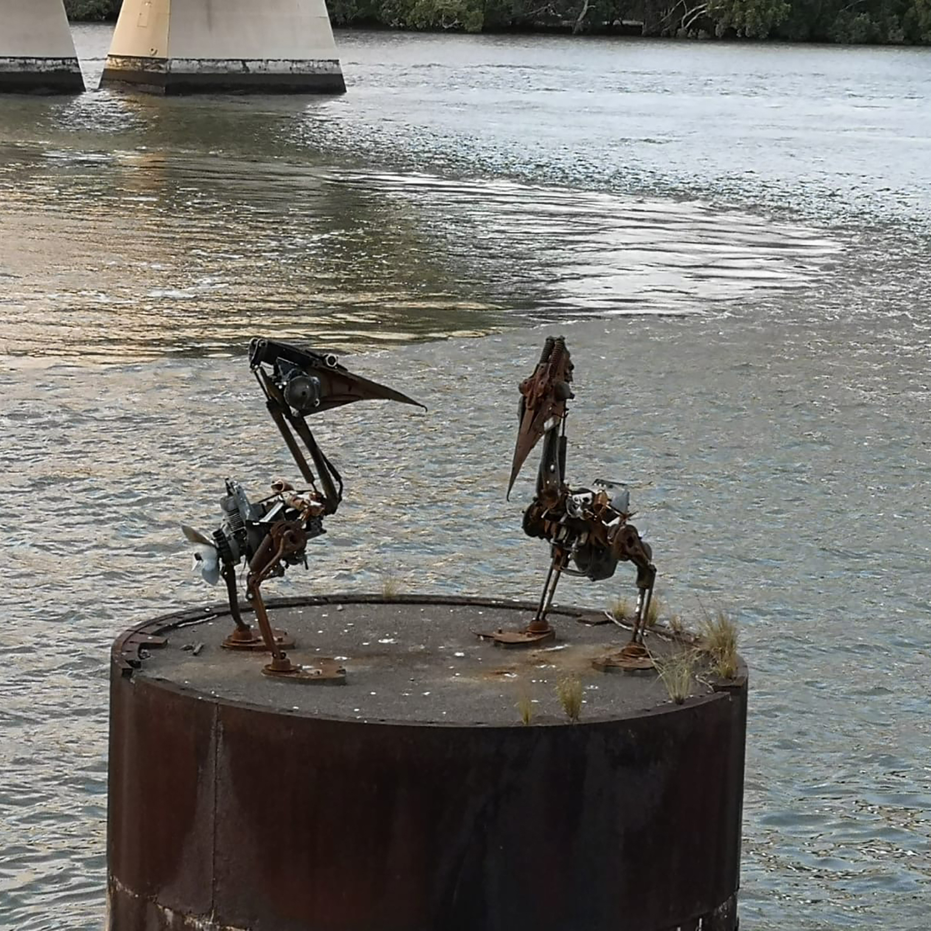 Zwei skulpturale Vögel aus Metallresten stehen auf einer rostigen Plattform im Wasser, mit einer Brücke im Hintergrund.
