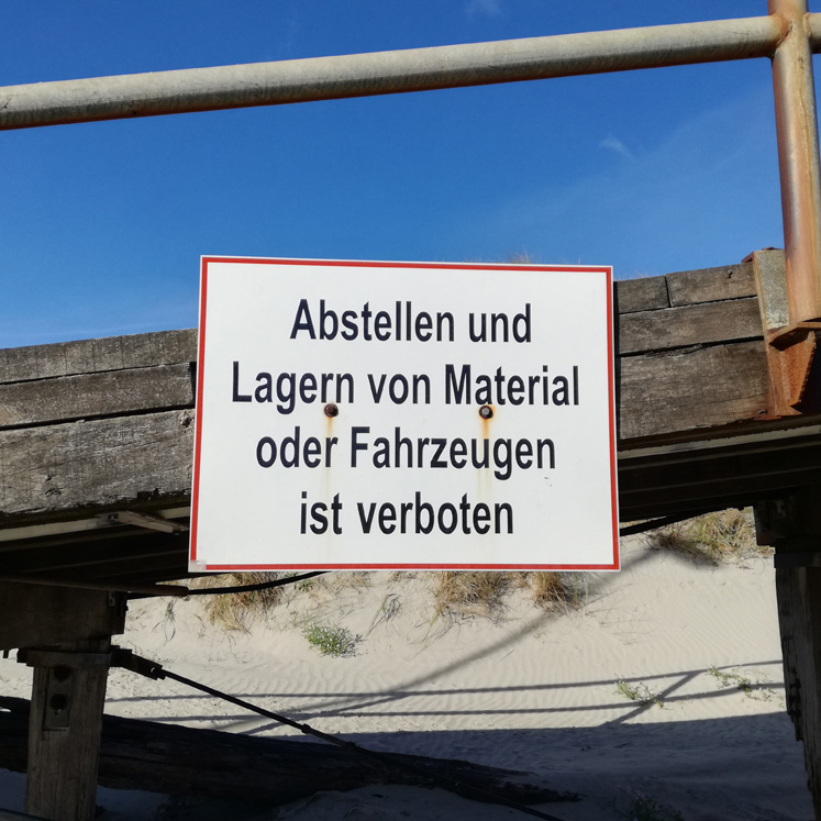 Ein Verbotsschild mit der Aufschrift „Abstellen und Lagern von Material oder Fahrzeugen ist verboten“ vor einer Holzwand mit blauem Himmel im Hintergrund