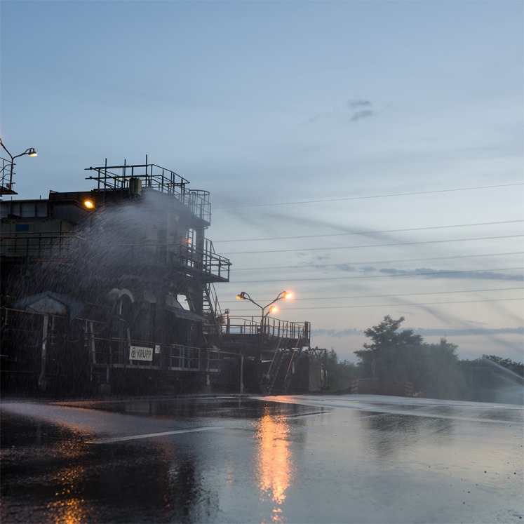 Eine Industrieanlage mit einem großen Rohrsystem wird mit Wasser besprüht. Die Szene ist in der Dämmerung aufgenommen, mit Lichtquellen an der Anlage