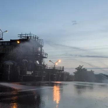 Eine Industrieanlage mit einem großen Rohrsystem wird mit Wasser besprüht. Die Szene ist in der Dämmerung aufgenommen, mit Lichtquellen an der Anlage