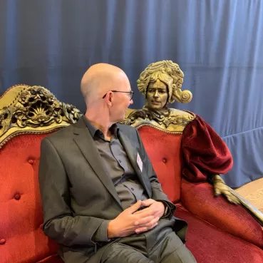 Ein Mann im Anzug sitzt auf einem roten Sofa und unterhält sich mit einer Statue