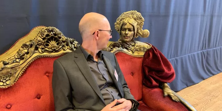 Ein Mann im Anzug sitzt auf einem roten Sofa und unterhält sich mit einer Statue