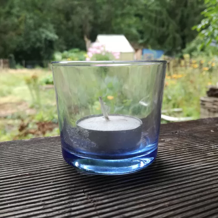 Ein blaues Glas mit einem Teelicht auf einer Holzfläche, im Hintergrund ein unscharfer Garten mit grünen Pflanzen