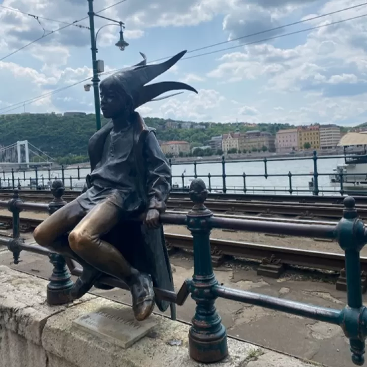 Die Statue der „Kleinen Prinzessin“ in Budapest, sitzend auf einem Geländer mit Blick auf die Donau und die Stadtlandschaft im Hintergrund