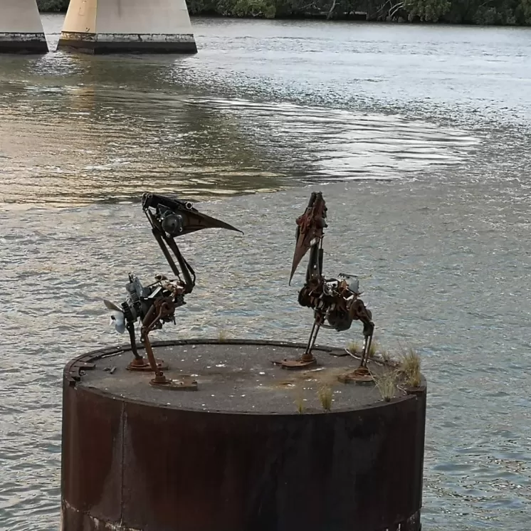 Zwei skulpturale Vögel aus Metallresten stehen auf einer rostigen Plattform im Wasser, mit einer Brücke im Hintergrund.