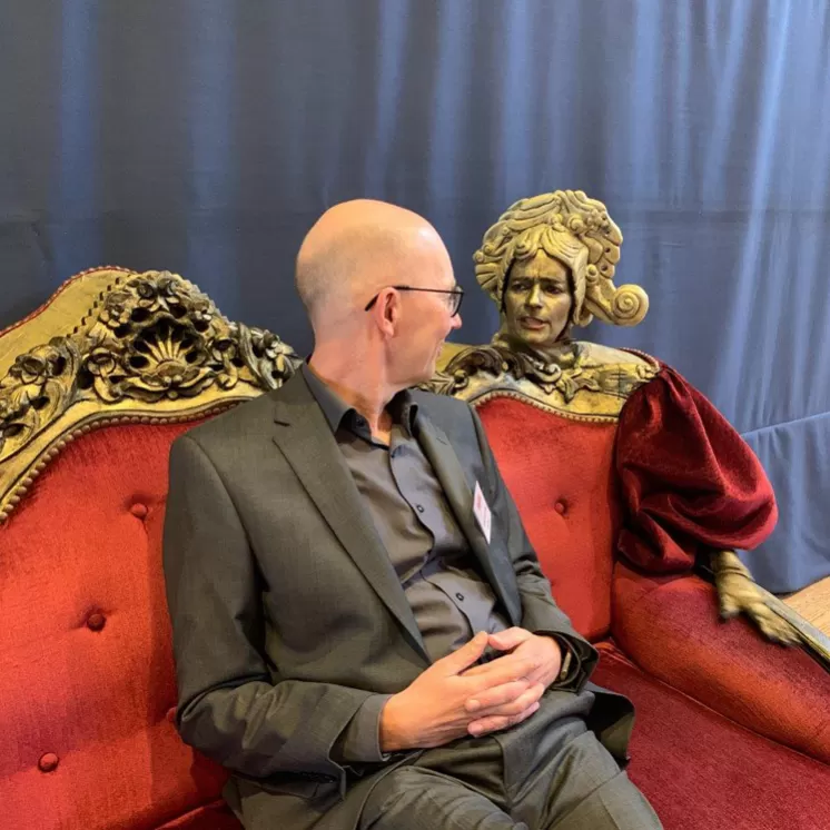 Ein Mann im Anzug sitzt auf einem reich verzierten roten Sofa und spricht mit einer als Statue verkleideten Person in barocker Kleidung.