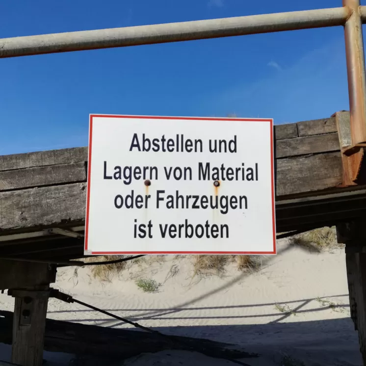 Ein Verbotsschild mit der Aufschrift „Abstellen und Lagern von Material oder Fahrzeugen ist verboten“ vor einer Holzwand mit blauem Himmel im Hintergrund