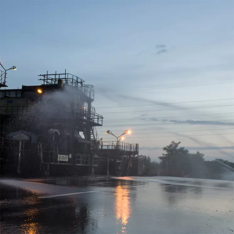 Eine Industrieanlage mit einem großen Rohrsystem wird mit Wasser besprüht. Die Szene ist in der Dämmerung aufgenommen, mit Lichtquellen an der Anlage